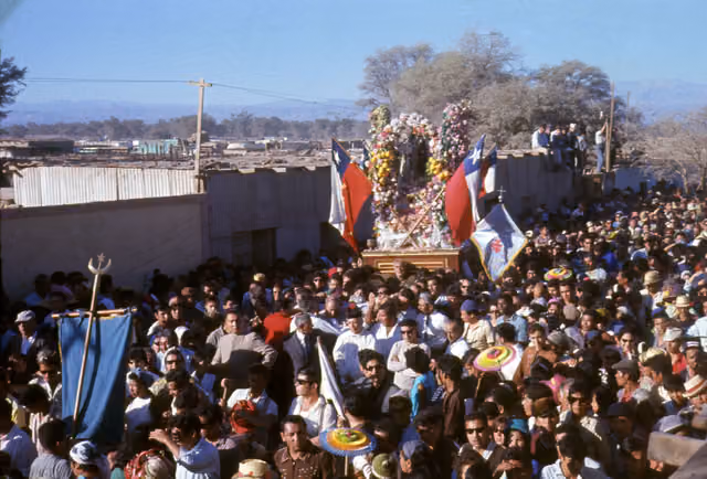 Fiesta de La Tirana: multitud con una gran corona de banderas en una plataforma, 1966-1967